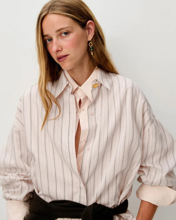 A woman wearing a pale blush pink button-up shirt with thin vertical dark stripes, styled with a black sweater and gold earrings against a light gray background.