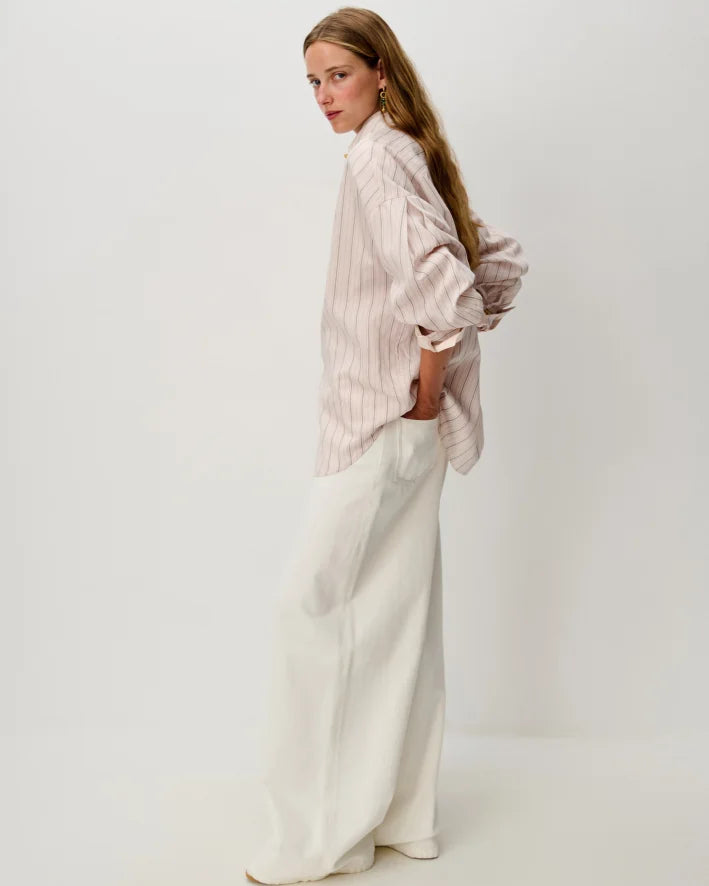 A woman stands in profile wearing a loose, pale pink and white vertically striped button-up shirt with long sleeves and white wide-leg trousers, photographed against a clean off-white studio background.