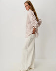 A woman stands in profile wearing a loose, pale pink and white vertically striped button-up shirt with long sleeves and white wide-leg trousers, photographed against a clean off-white studio background.