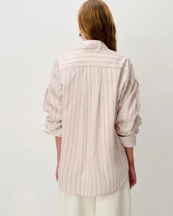 A woman shown from the back wearing a light pink and beige vertically striped button-up shirt with rolled-up sleeves, paired with white trousers, against a plain light gray background.