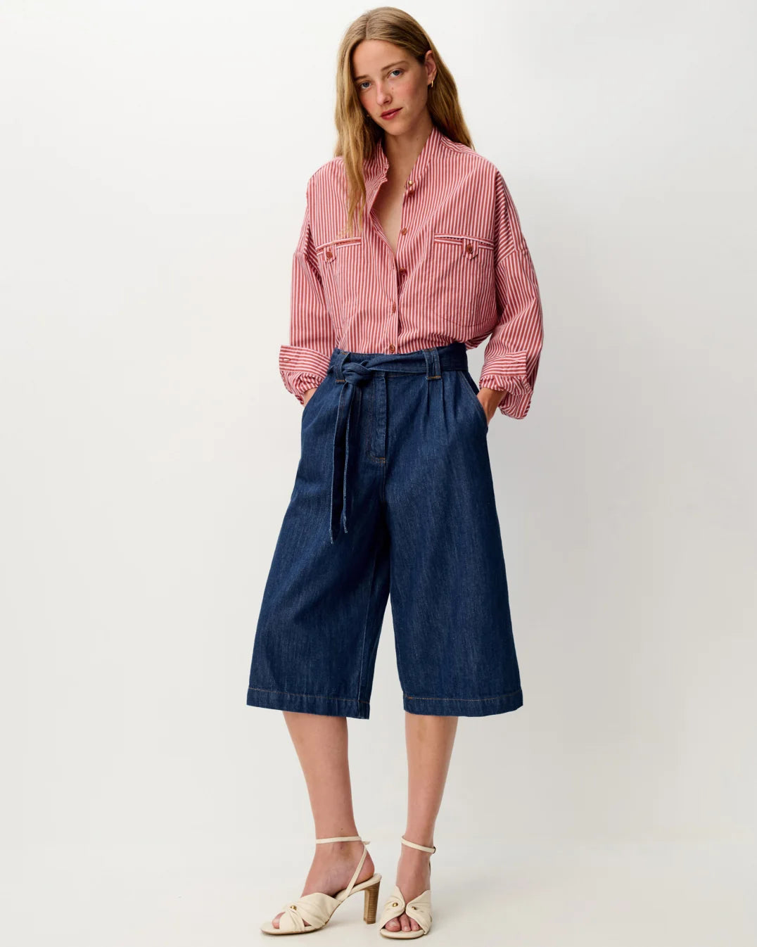 A woman modeling a pink and white striped button-down shirt tucked into dark blue denim culottes with a tie waist, paired with beige heeled sandals, against a plain light gray studio background.