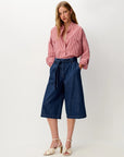 A woman modeling a pink and white striped button-down shirt tucked into dark blue denim culottes with a tie waist, paired with beige heeled sandals, against a plain light gray studio background.