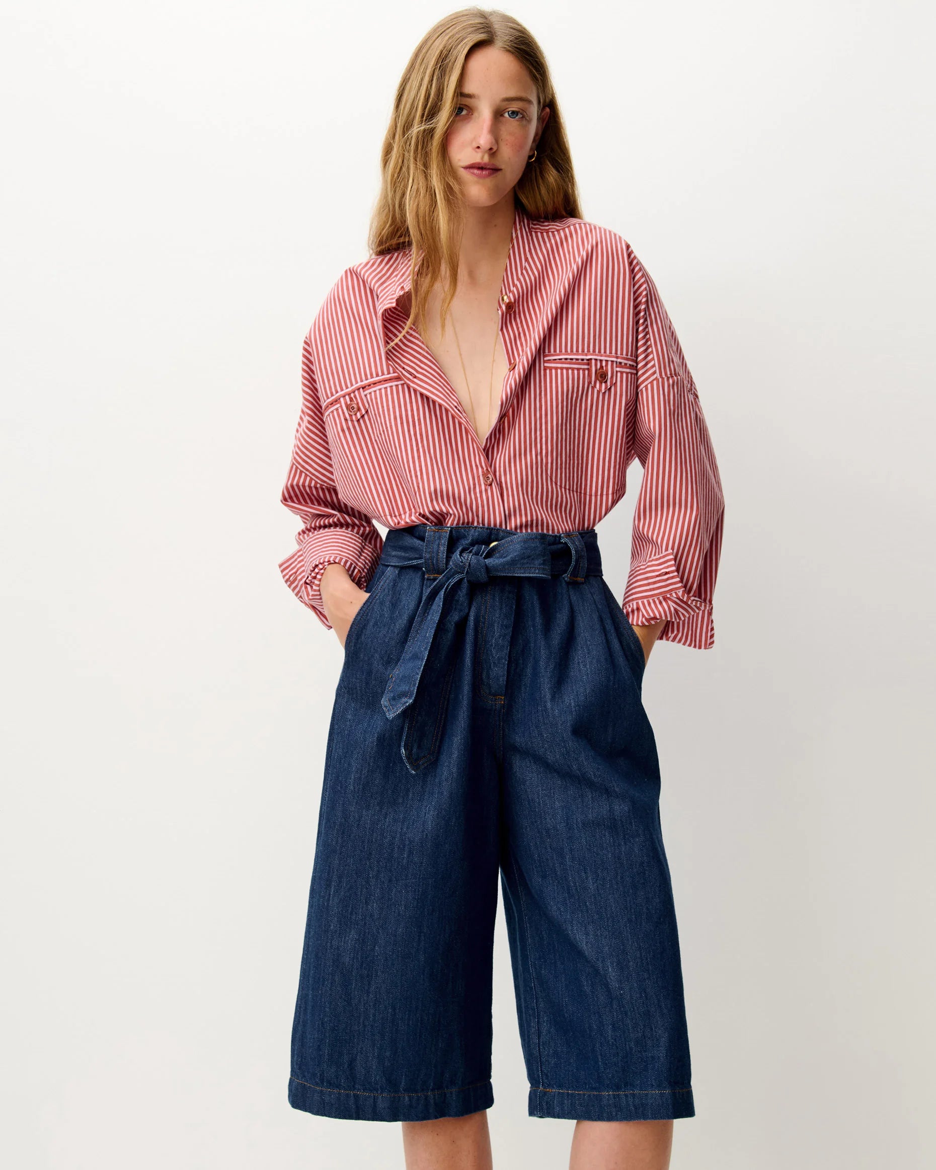 A woman modeling a pink and white vertically striped button-down shirt paired with dark blue denim culottes featuring a matching waist tie, set against a plain off-white studio background.