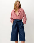 A woman modeling a pink and white vertically striped button-down shirt paired with dark blue denim culottes featuring a matching waist tie, set against a plain off-white studio background.