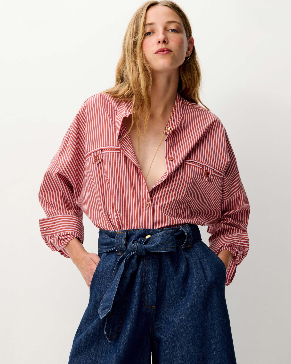 A woman wearing a pink and white vertically striped button-up shirt with chest pockets, tucked into dark blue high-waisted wide-leg jeans with a tie waist, against a plain light gray background.