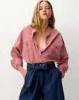 A woman wearing a pink and white vertically striped button-up shirt with chest pockets, tucked into dark blue high-waisted wide-leg jeans with a tie waist, against a plain light gray background.