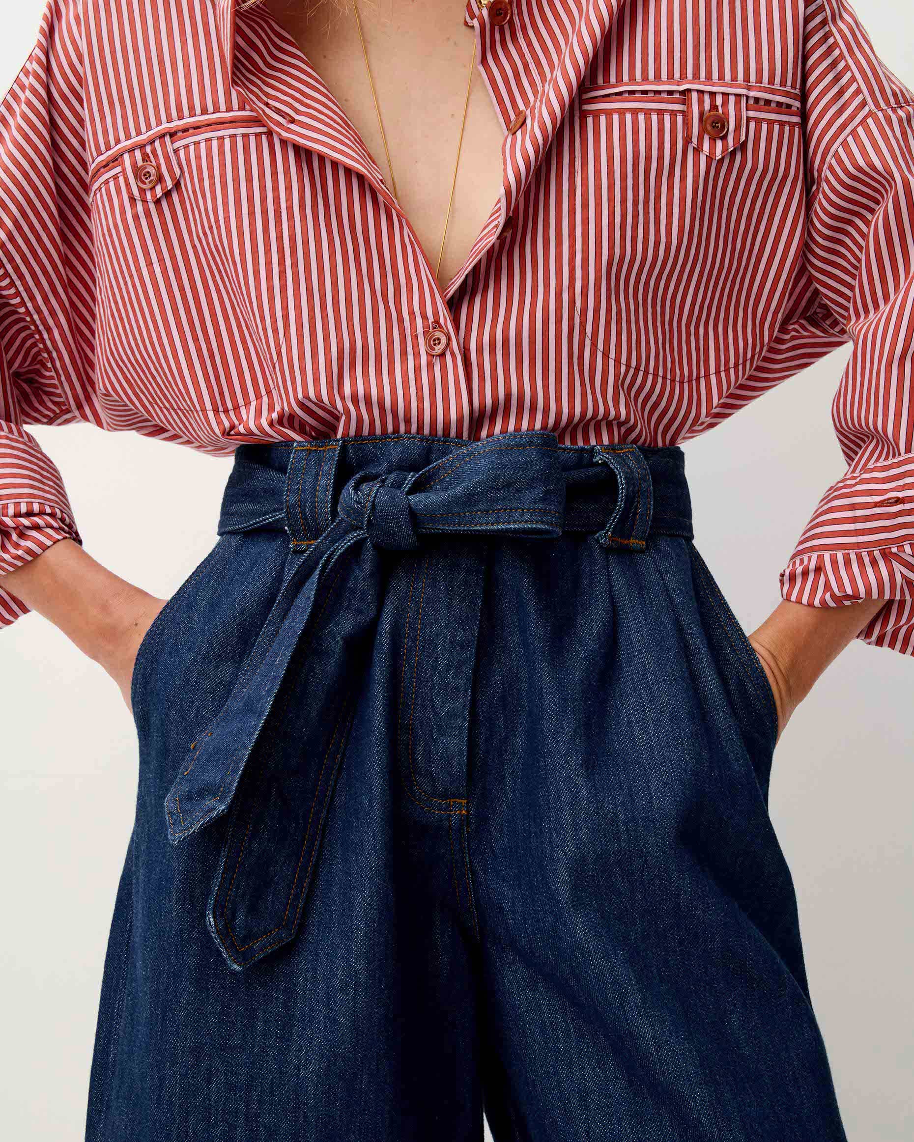 A close-up of a person wearing a red and white vertically striped button-up shirt and dark blue high-waisted denim culottes with a matching tie belt.