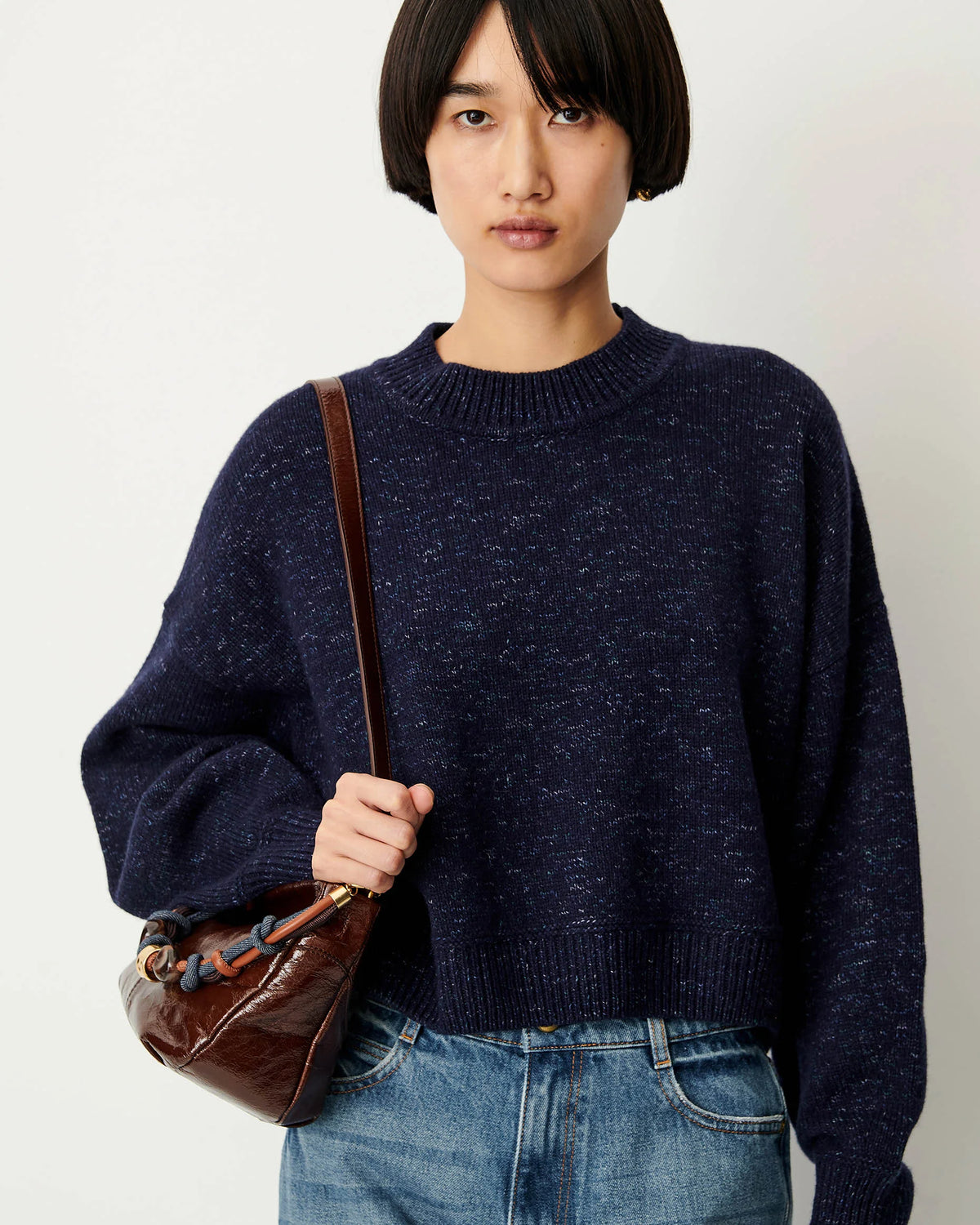 A model with a short dark bob haircut wearing an oversized navy blue heathered knit sweater and blue jeans, carrying a brown leather shoulder bag against a plain off-white background.