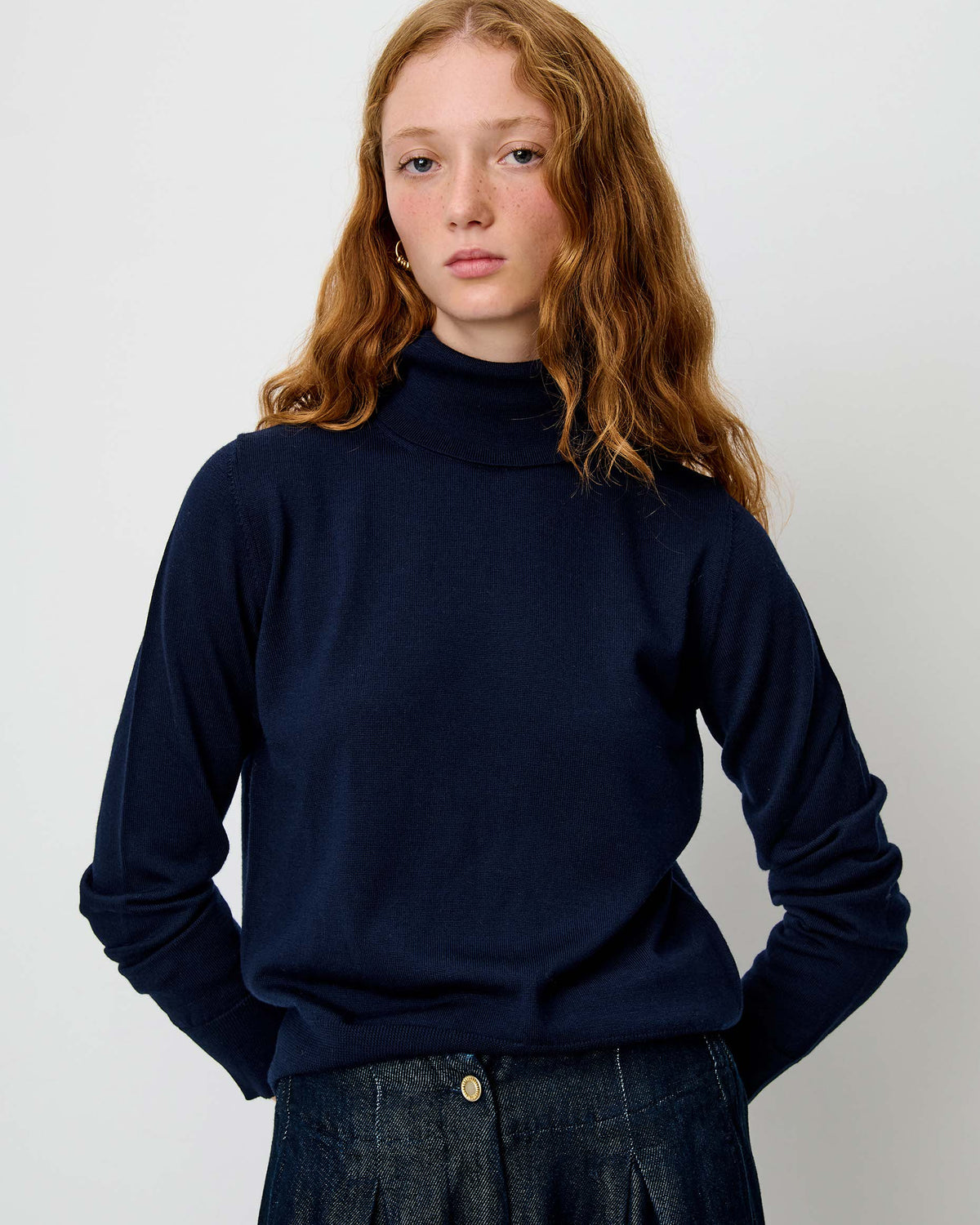 A young woman with wavy red hair wearing a dark navy blue turtleneck sweater and dark denim trousers, standing against a plain light gray background.