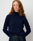 A young woman with wavy red hair wearing a dark navy blue turtleneck sweater and dark denim trousers, standing against a plain light gray background.