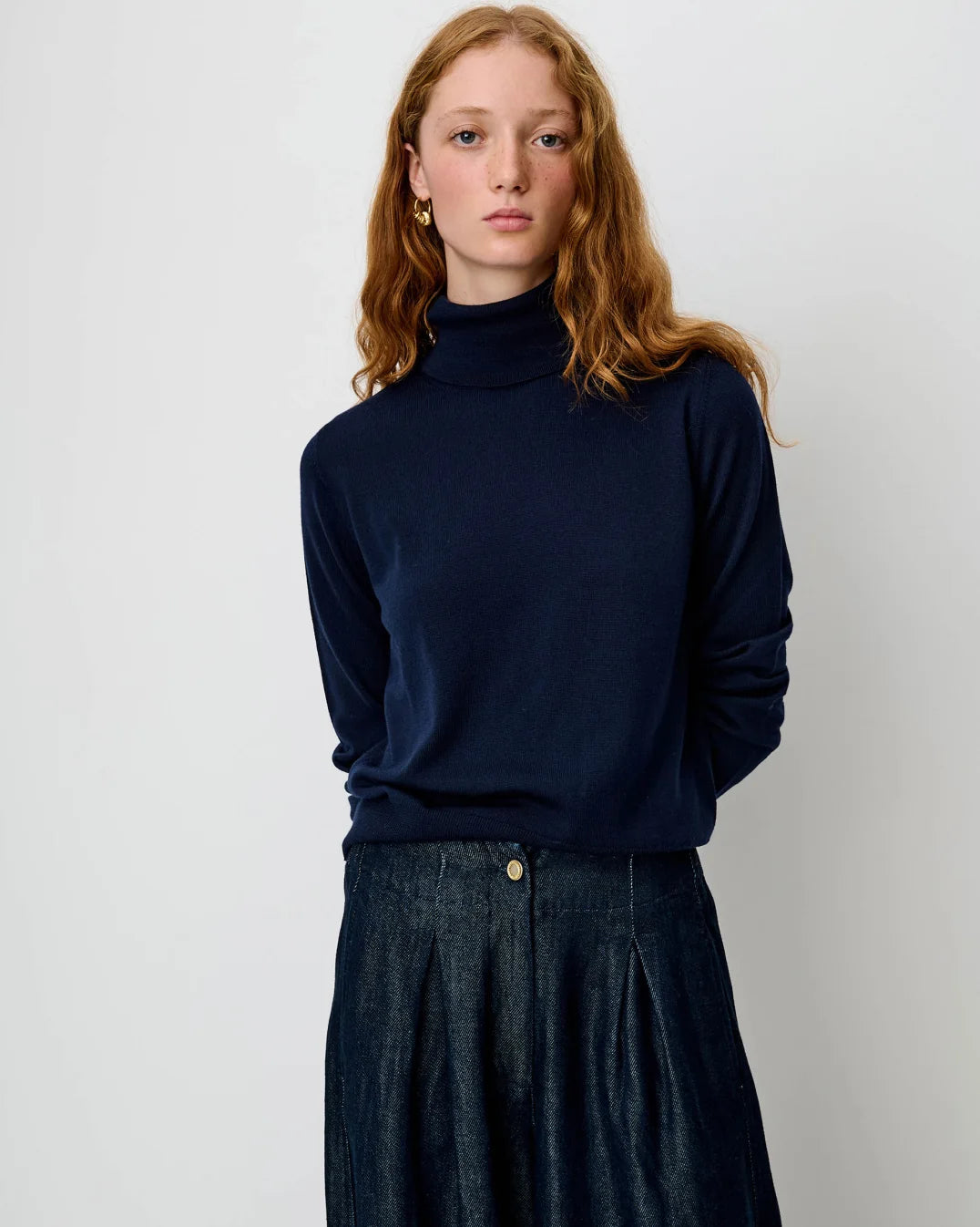 A woman with red hair wearing a dark navy blue ribbed turtleneck sweater and a dark denim skirt, standing against a plain light gray background.