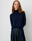 A woman with red hair wearing a dark navy blue ribbed turtleneck sweater and a dark denim skirt, standing against a plain light gray background.