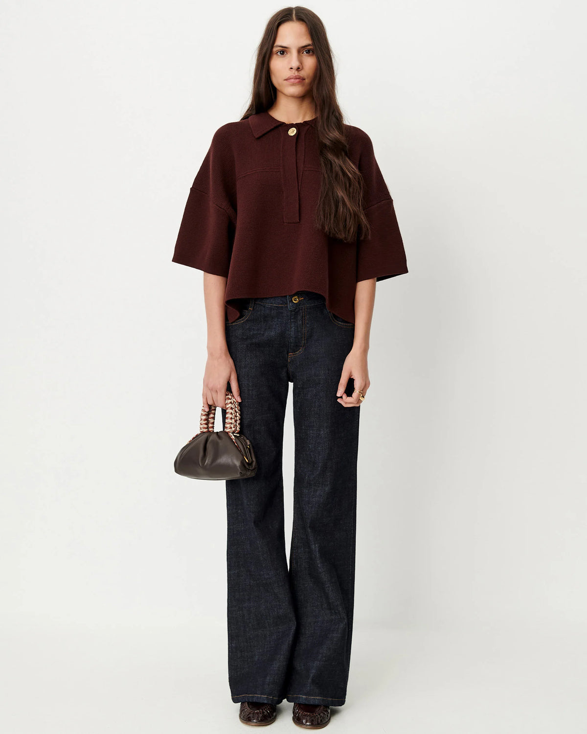 A woman stands against a plain light gray studio background wearing a cropped short-sleeve collared top in deep burgundy, dark wide-leg jeans, and holding a small dark brown handbag.