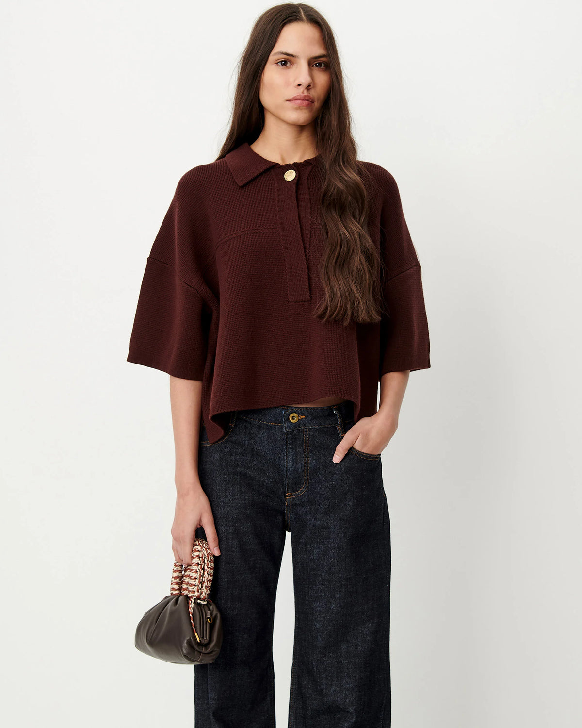 A woman modeling a dark brown short-sleeve knit polo-style top with a button placket, paired with dark blue jeans and holding a small dark handbag, against a plain light gray studio background.