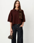 A woman modeling a dark brown short-sleeve knit polo-style top with a button placket, paired with dark blue jeans and holding a small dark handbag, against a plain light gray studio background.