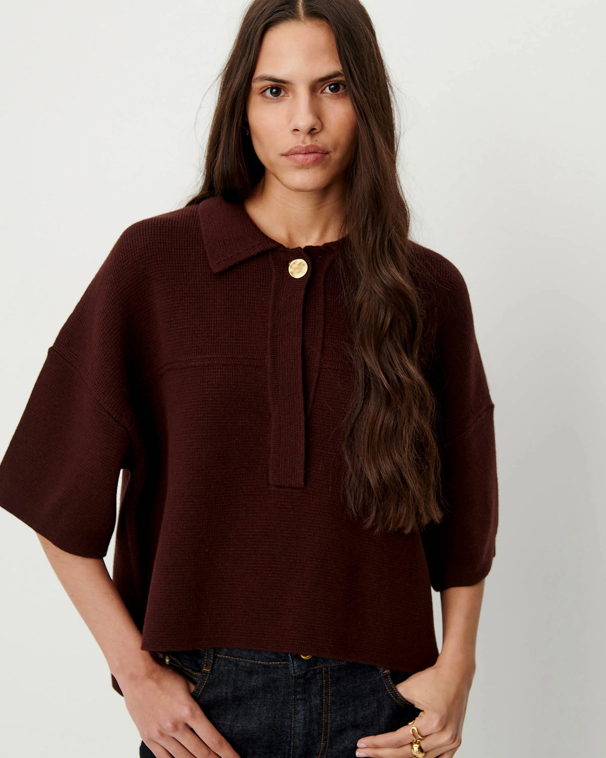 A woman wearing a dark burgundy knit polo sweater with a single gold button closure at the collar, paired with dark blue jeans, against a plain light gray background.