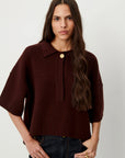 A woman wearing a dark burgundy knit polo sweater with a single gold button closure at the collar, paired with dark blue jeans, against a plain light gray background.