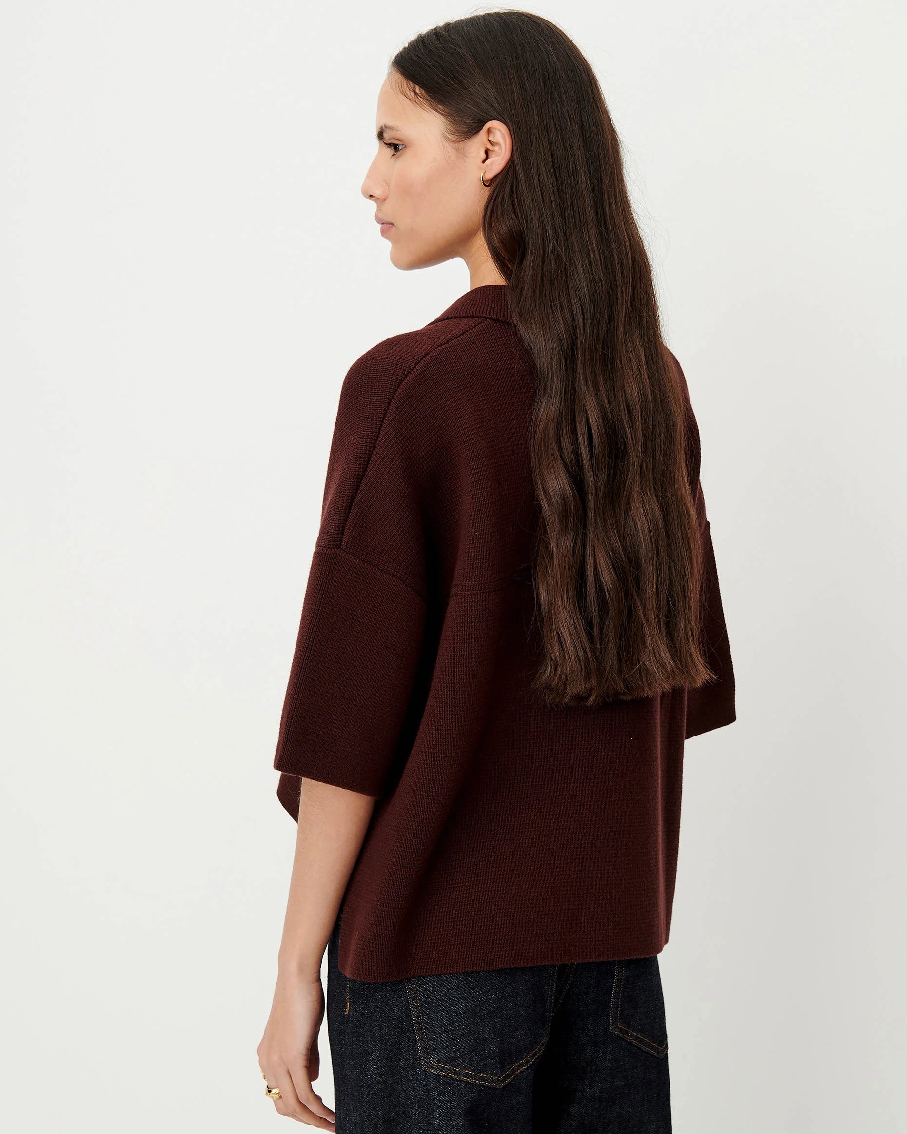 A woman with long dark brown hair is shown from a side-back angle wearing a dark brown knit sweater and dark blue jeans against a plain white studio background.