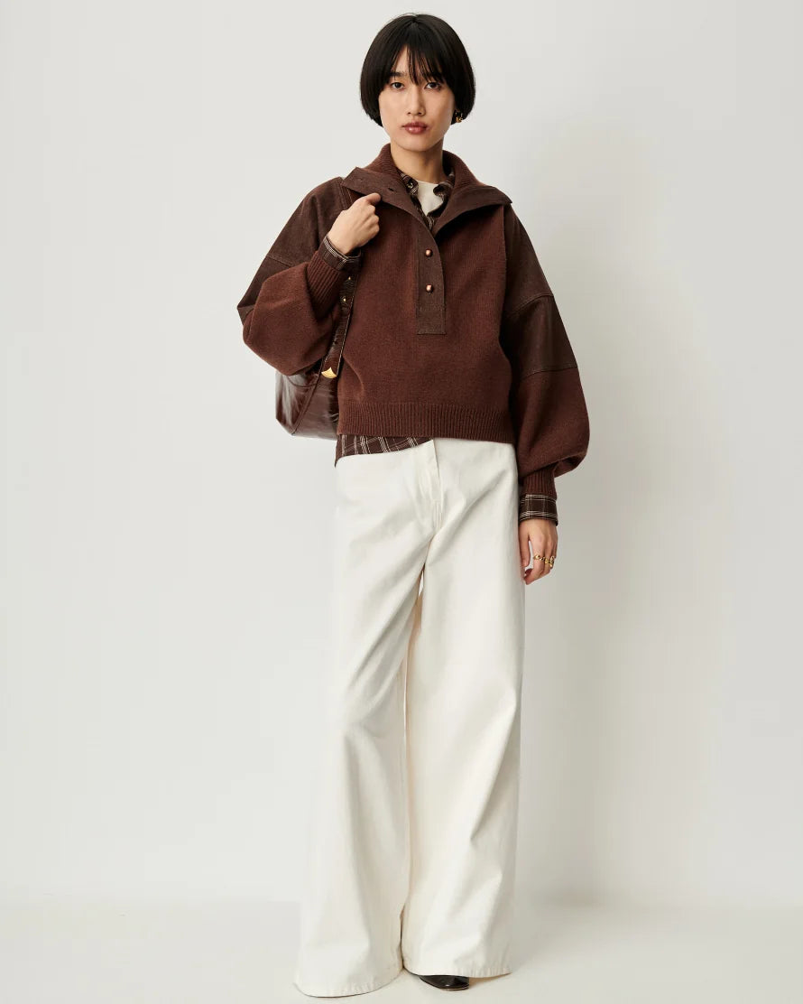 A model wearing a brown knit mock-neck sweater with textured sleeves, paired with wide-leg cream trousers and a brown shoulder bag, standing against a plain light gray background.