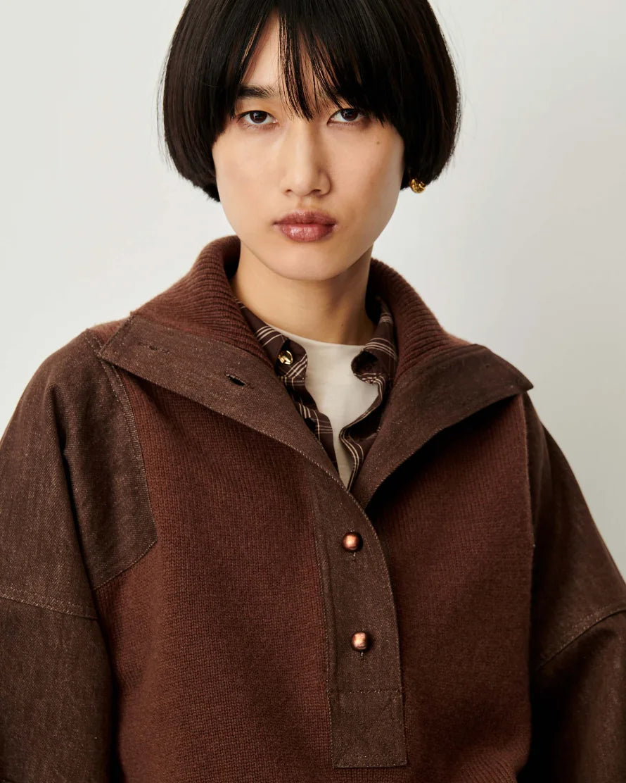 A model with a short dark bob haircut wearing a brown corduroy jacket with a high collar and button details over a patterned shirt and white undershirt, set against a plain light gray background.