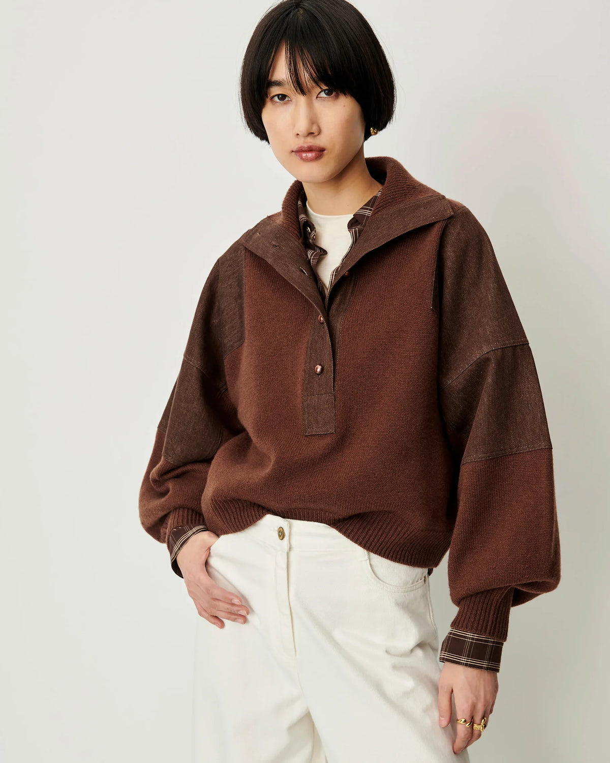 A model wearing a brown color-blocked knit pullover sweater with a high collar and white pants, photographed against a light gray studio background.