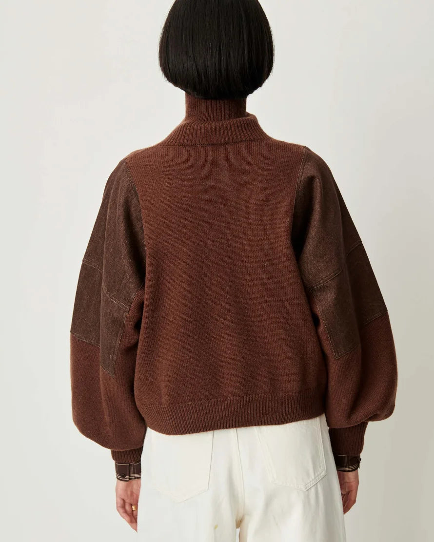 A person shown from the back wearing a brown knit sweater with darker brown patchwork sleeves and white pants, photographed against a plain light background.
