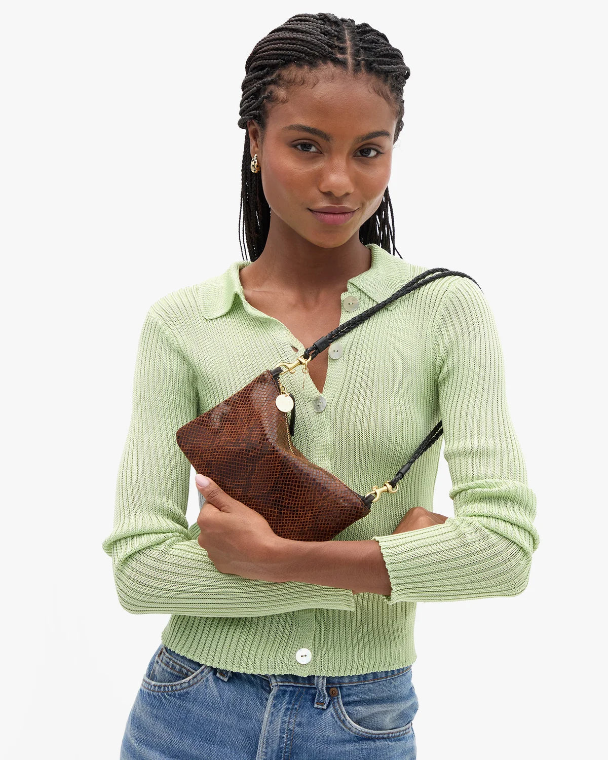 A woman wearing a light green ribbed knit polo cardigan and blue jeans holds a brown textured leather shoulder bag with a black strap and gold hardware against a plain white background.