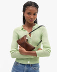 A woman wearing a light green ribbed knit polo cardigan and blue jeans holds a brown textured leather shoulder bag with a black strap and gold hardware against a plain white background.