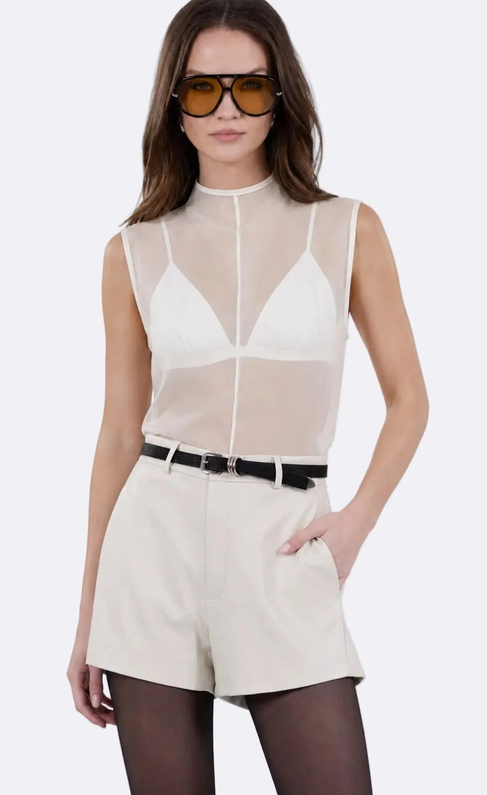 A model wearing a sleeveless white mesh mock-neck top over a white bralette, paired with Robbie Short and a black belt, against a light gray studio background.