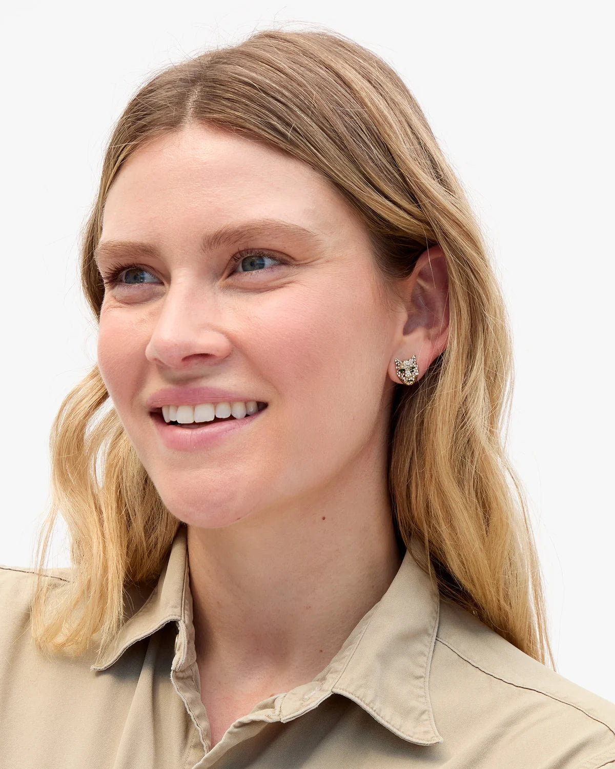 A smiling woman with blonde hair wearing a beige collared shirt and a small silver-toned stud earring with a floral design, photographed against a clean white background.