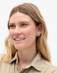 A smiling woman with blonde hair wearing a beige collared shirt and a small silver-toned stud earring with a floral design, photographed against a clean white background.