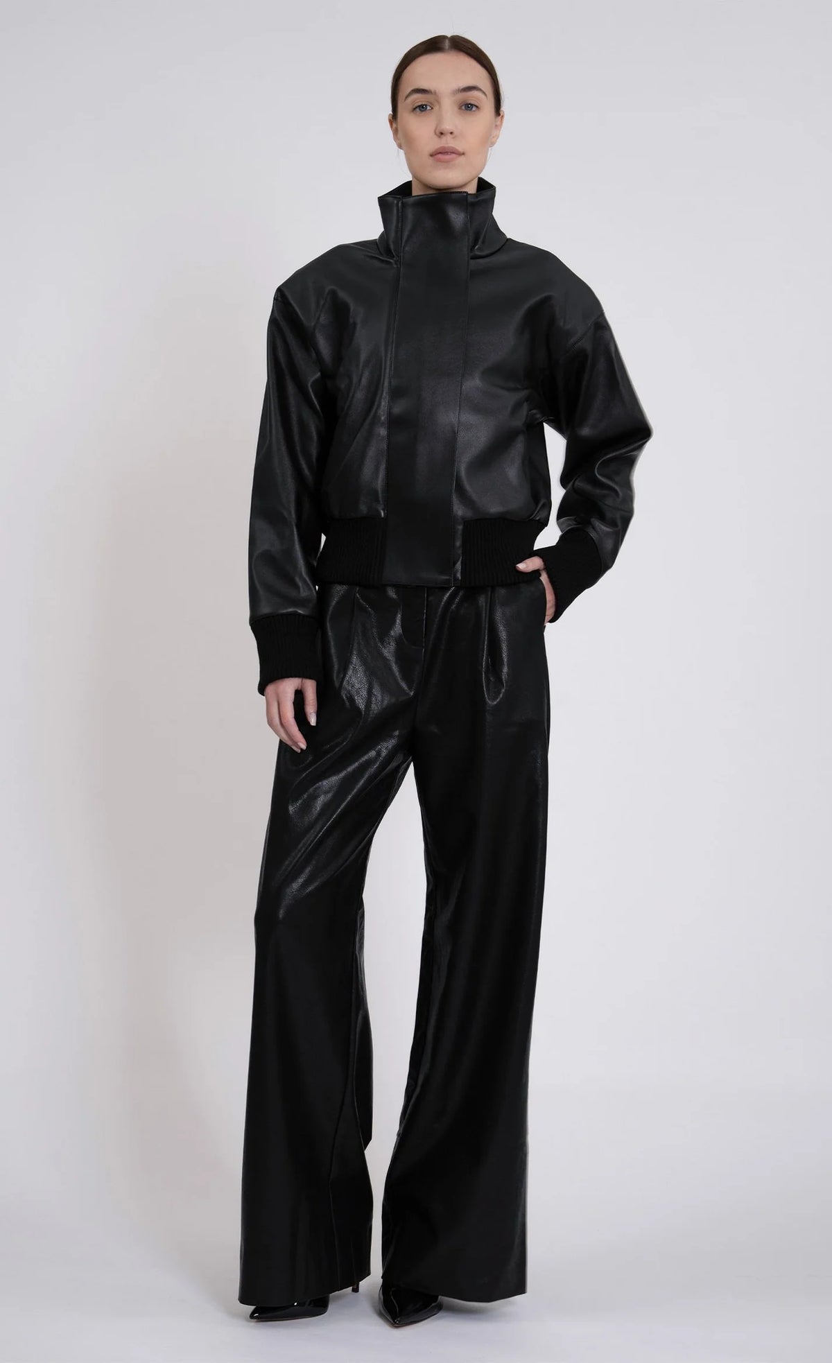 A model wearing a black faux-leather two-piece outfit consisting of a cropped high-collar jacket and matching wide-leg trousers, photographed against a light gray studio background.
