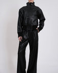 A model wearing a black faux-leather two-piece outfit consisting of a cropped high-collar jacket and matching wide-leg trousers, photographed against a light gray studio background.