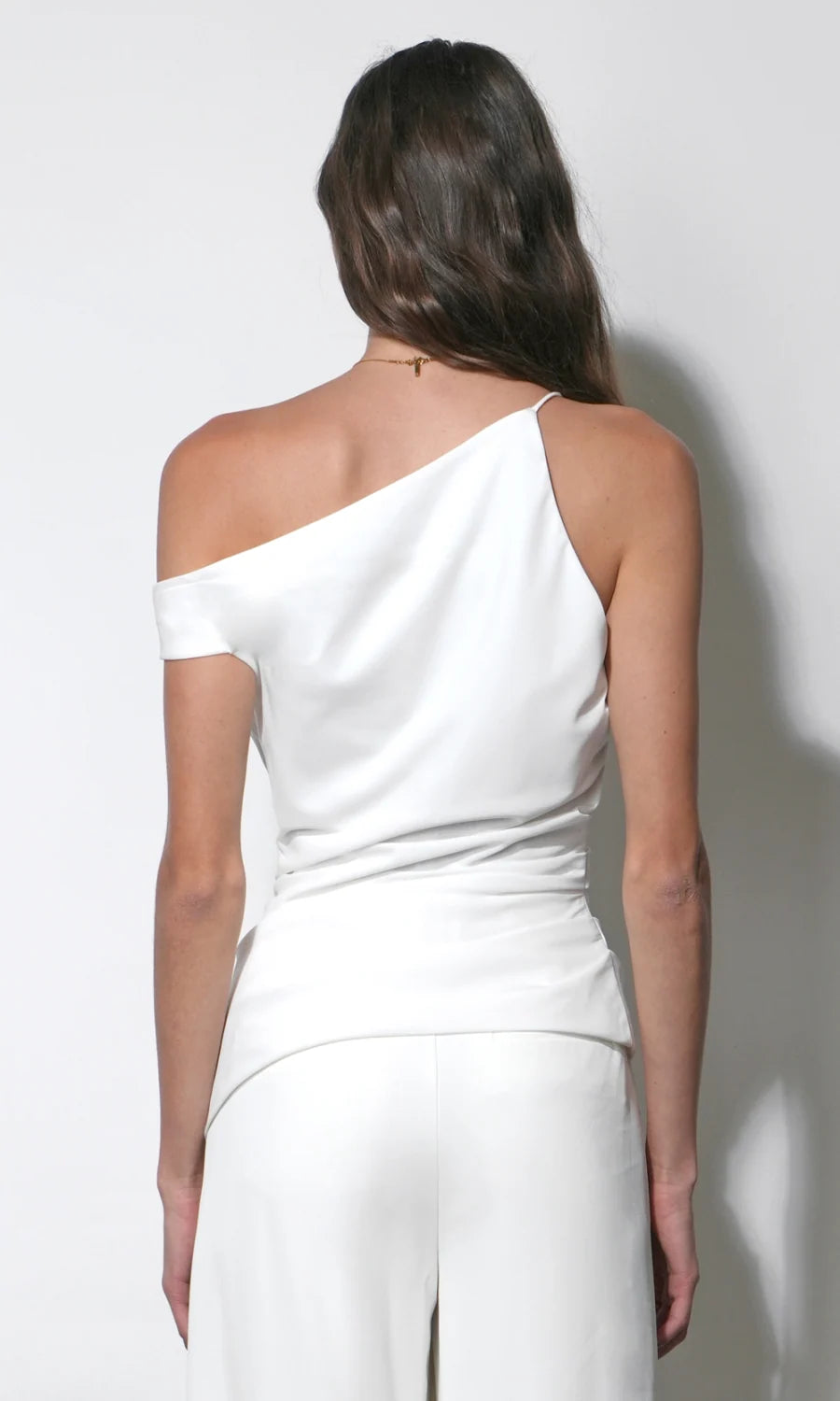 A woman shown from the back wearing a white one-shoulder top and matching white pants against a plain light gray studio background.