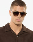 A man wearing aviator-style sunglasses with dark brown lenses and thin gold-tone metal frames, paired with a brown textured collared shirt against a plain white background.