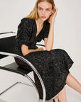 Woman in a black patterned dress sitting on a modern chair against a white background