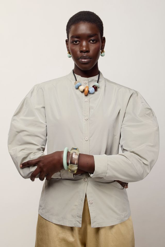 A model wearing a light beige, oversized long-sleeve button-up shirt with voluminous balloon sleeves, paired with beige trousers and colorful statement jewelry, against a plain light gray background.