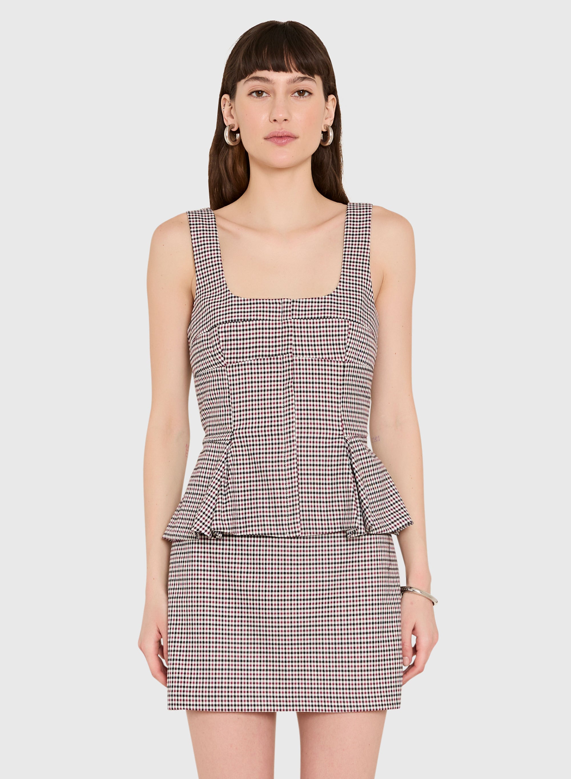 A woman modeling a sleeveless mini set with a square neckline and a peplum waist, featuring a fine brown, beige, and white gingham pattern, set against a solid light gray background.
