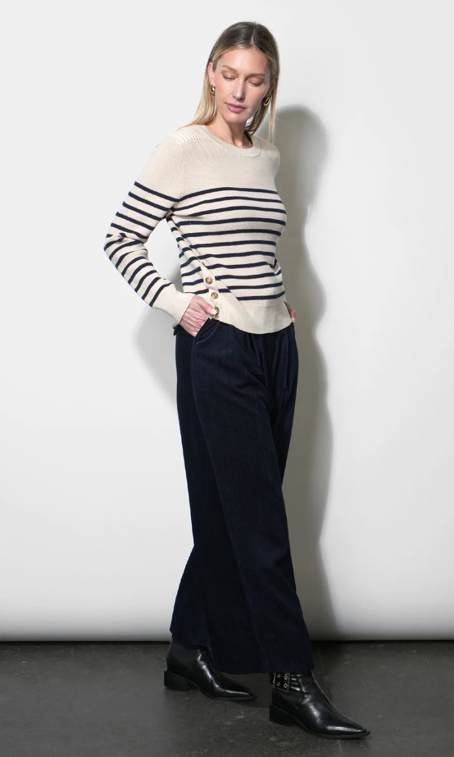 A woman modeling a cream knit sweater with black horizontal stripes on the sleeves and upper chest, paired with dark wide-leg trousers and black ankle boots, standing against a plain light gray wall.