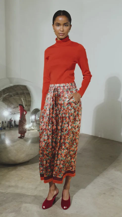 A model stands in a gallery wearing a bright red long-sleeve turtleneck sweater and a high-waisted, pleated midi skirt with a dense red, orange, and green floral pattern. She is wearing red pointed-toe flats.