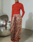 A model stands in a gallery wearing a bright red long-sleeve turtleneck sweater and a high-waisted, pleated midi skirt with a dense red, orange, and green floral pattern. She is wearing red pointed-toe flats.