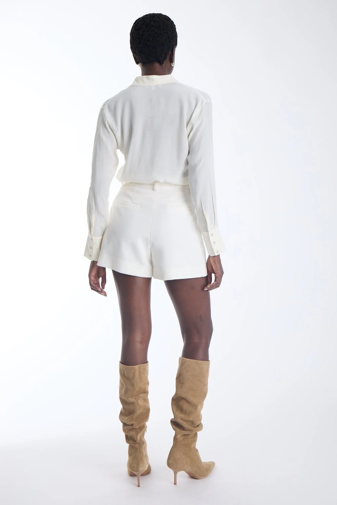 A model shown from the back wearing a white long-sleeve collared shirt, white shorts, and tan knee-high suede boots against a plain white studio background.