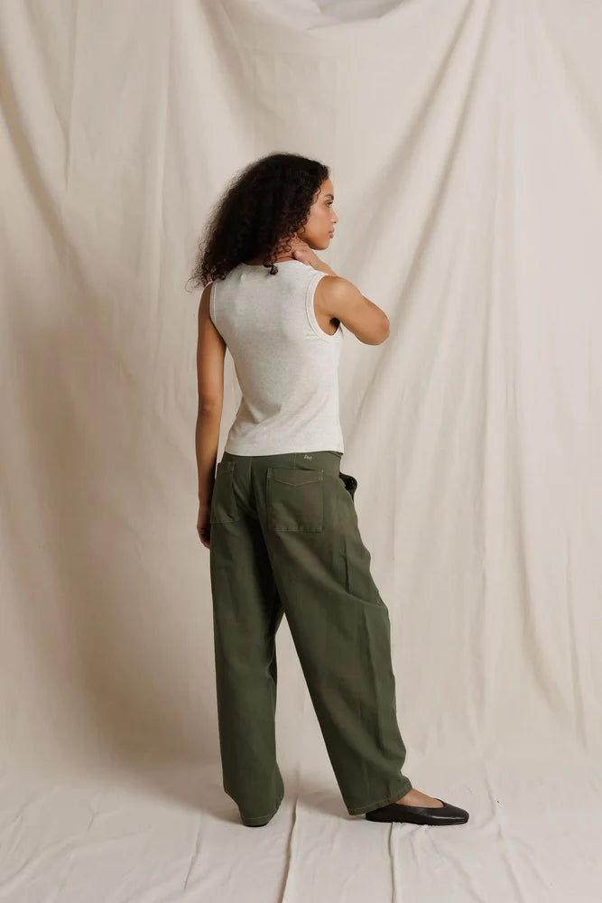 A woman stands in profile wearing wide-leg olive green trousers, a white ribbed tank top, and black flats, posed against a draped off-white fabric backdrop.