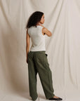 A woman stands in profile wearing wide-leg olive green trousers, a white ribbed tank top, and black flats, posed against a draped off-white fabric backdrop.