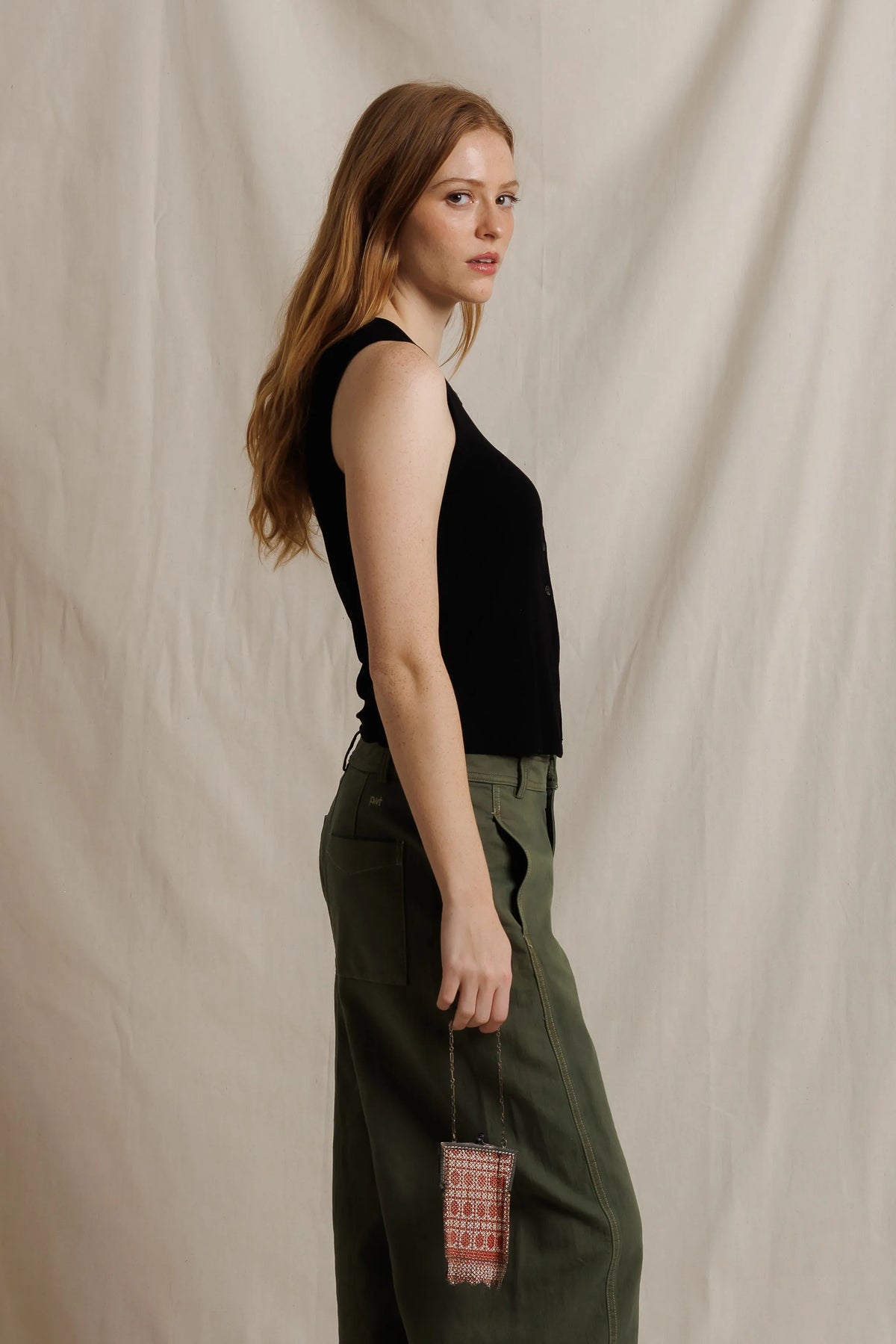 model wearing a sleeveless button down black top with green pants on a cream background, side view