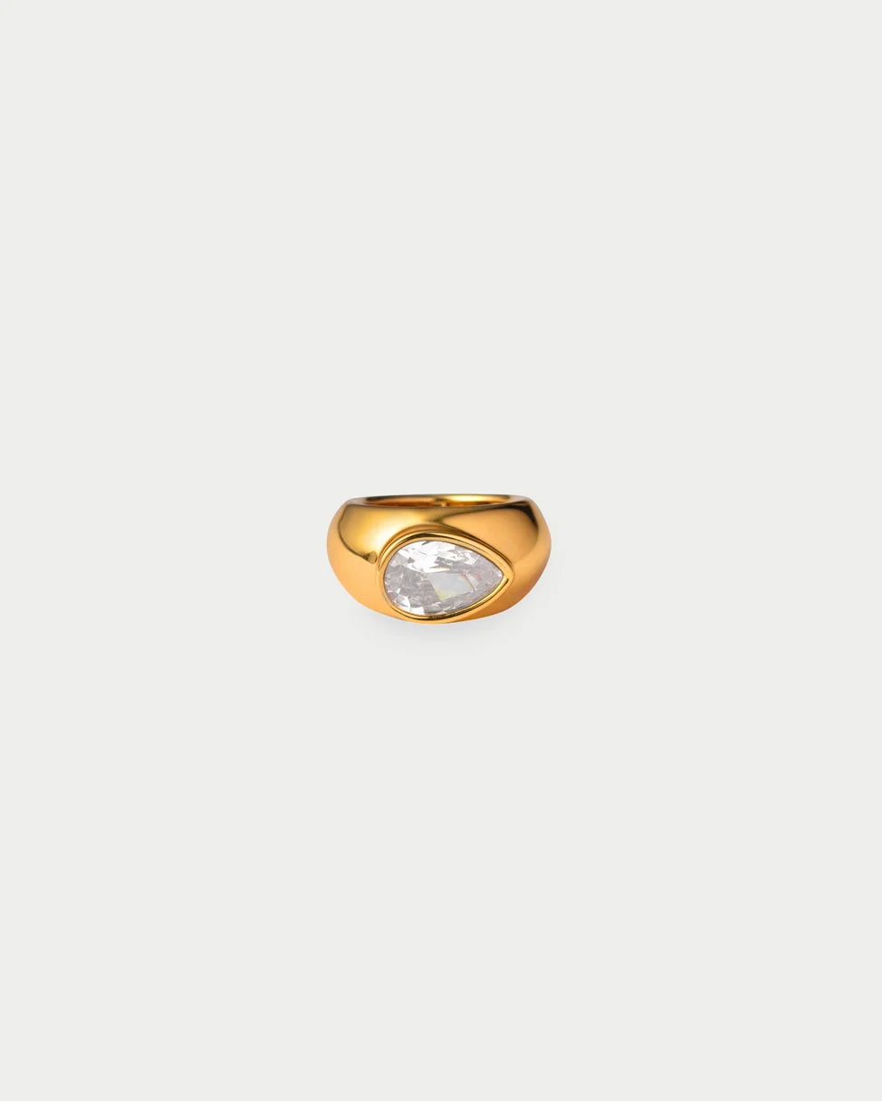 A gold ring featuring a teardrop-shaped clear gemstone, set against a solid light gray background.