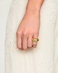 A close-up of a person's hand wearing a gold ring with a clear oval gemstone, set against a cream-colored embroidered dress.