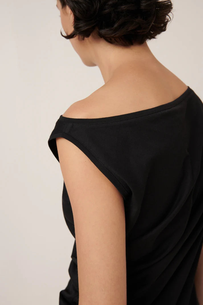 asymmetric top in black on a model with green pants on a cream background, shoulder view