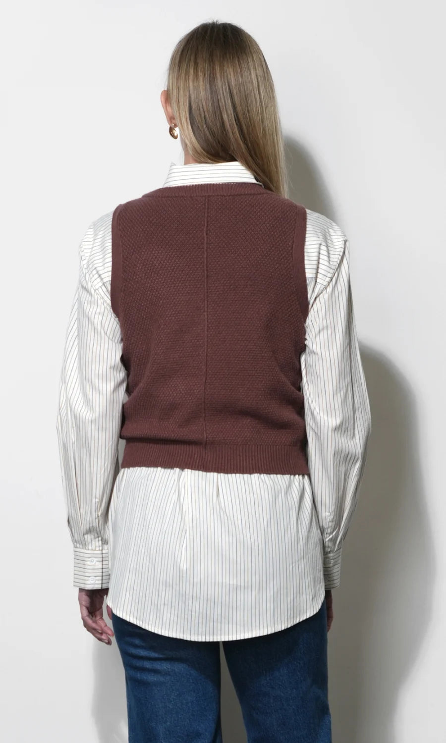 A woman shown from the back wearing a brown ribbed knit sweater vest over a white and gray vertically striped button-down shirt, paired with blue jeans, against a plain light gray wall.