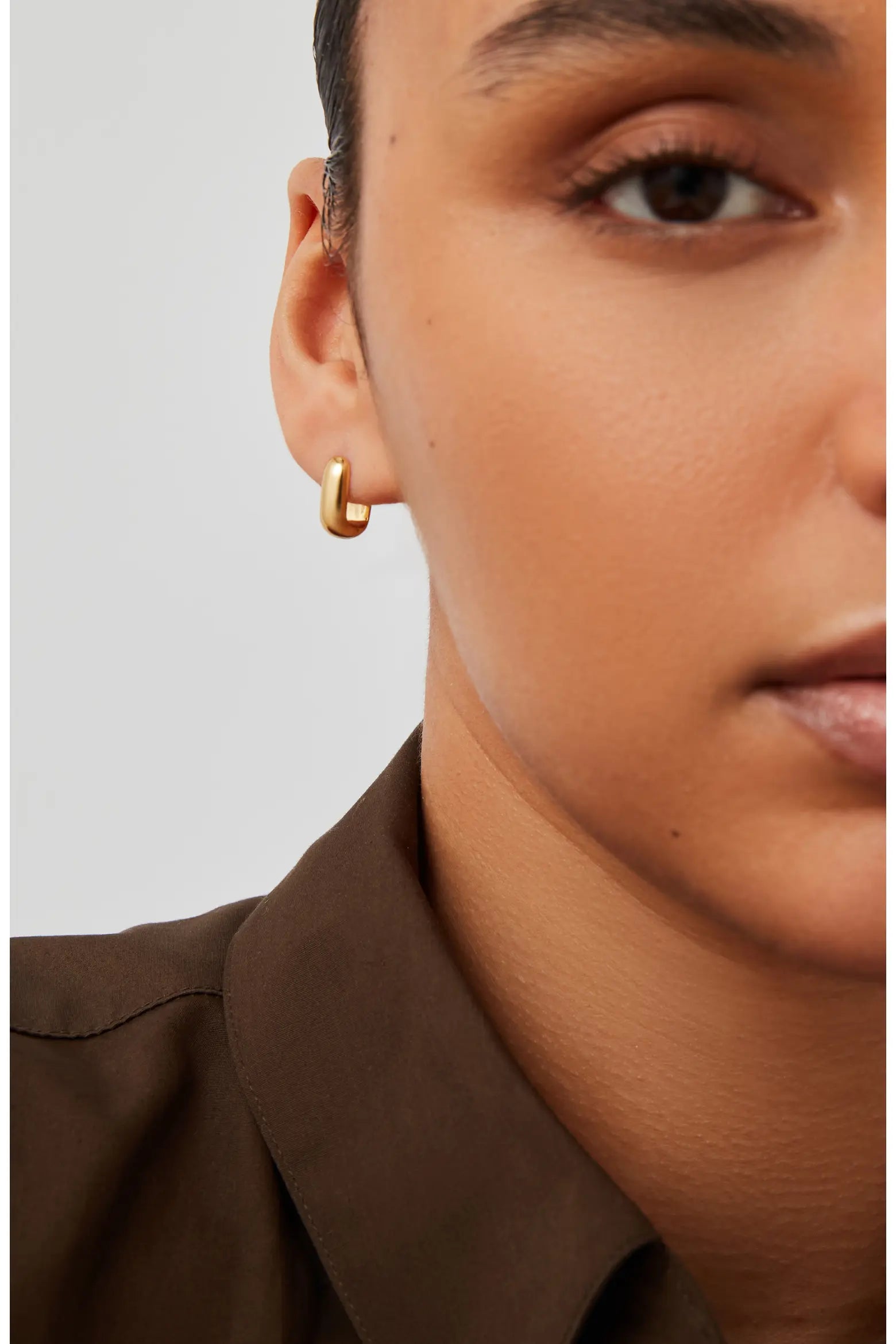 A close-up of a model wearing a small, polished gold huggie hoop earring, paired with a dark brown collared shirt against a light gray background.